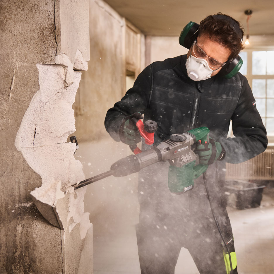 Bărbat cu ciocan demolator, mască și căști de protecție, demolând un perete.