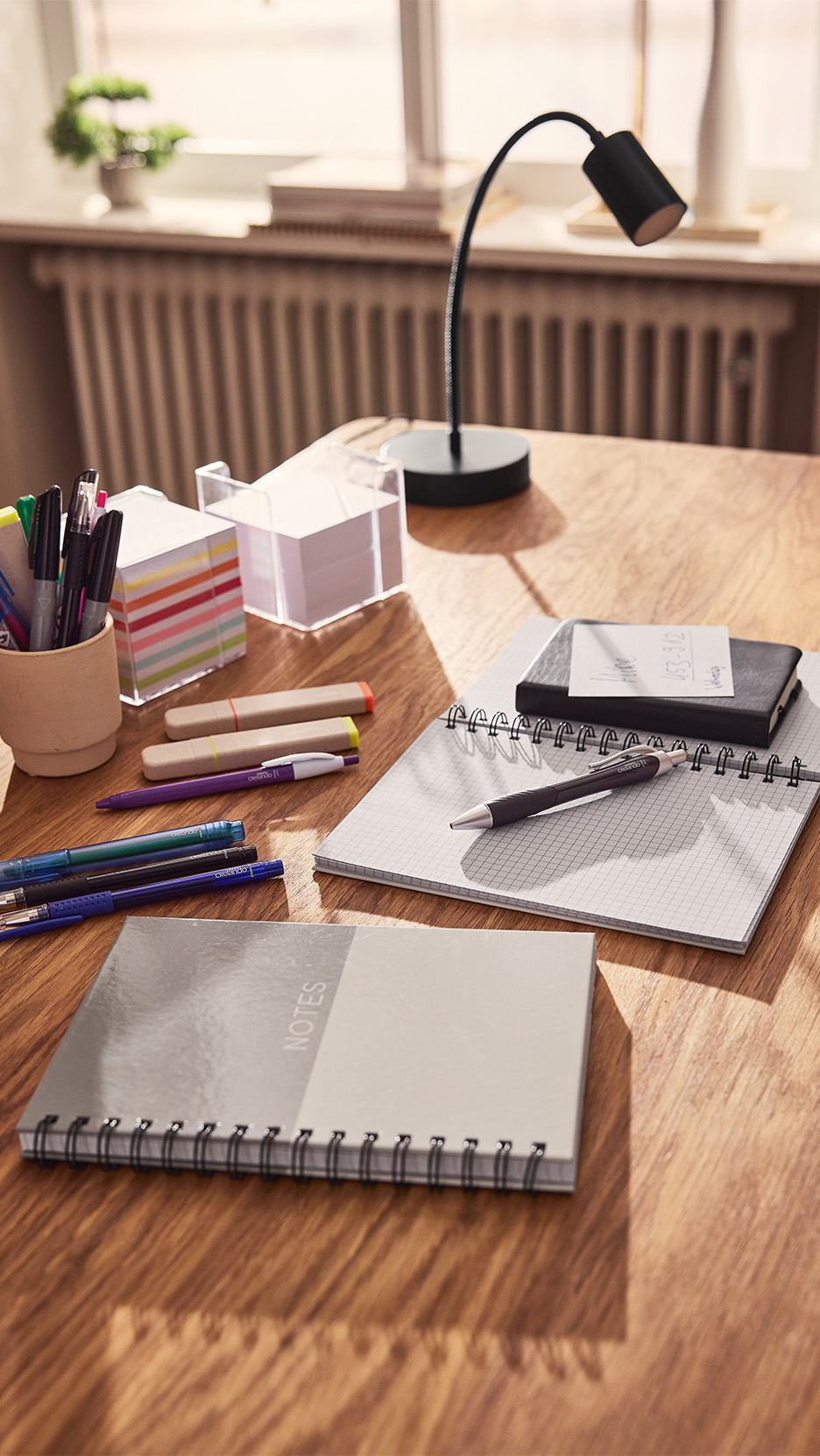 Birou din lemn cu caiete, pixuri, markere și o lampă de birou.