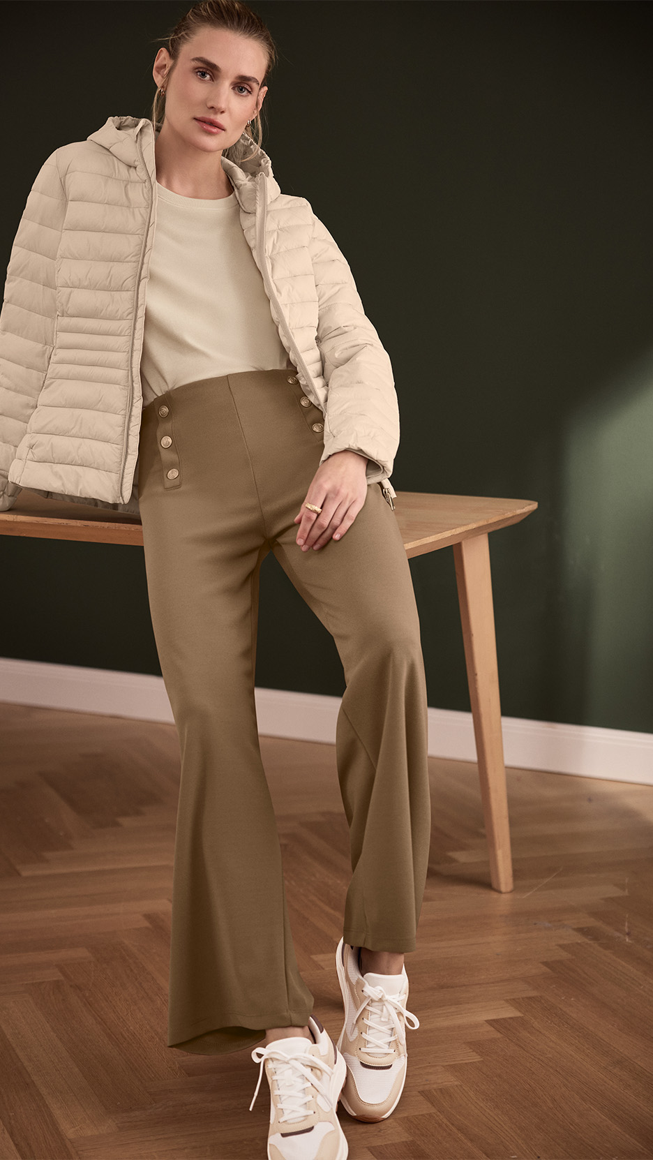 Femeie într-o geacă bej matlasată, pantaloni maro cu talie înaltă și adidași albi, sprijinită de o masă.