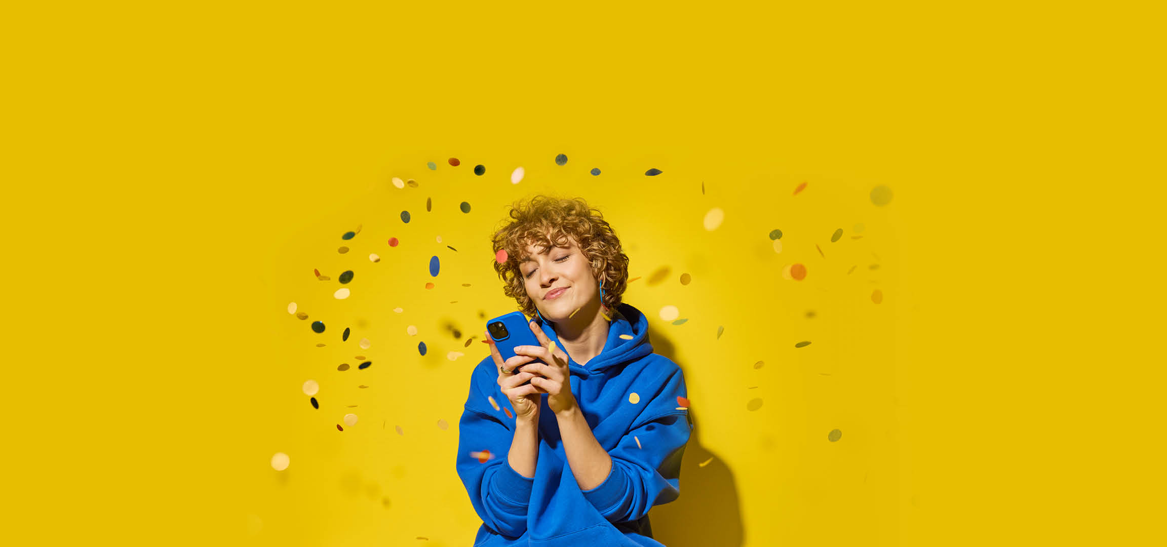 Femeie zâmbitoare în hanorac albastru, cu telefonul în mână, înconjurată de confetti pe fundal galben.