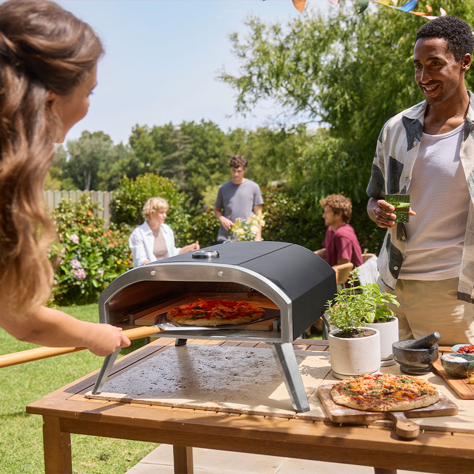 Familie la o petrecere în grădină, femeie punând pizza într-un cuptor, bărbat ținând o băutură.