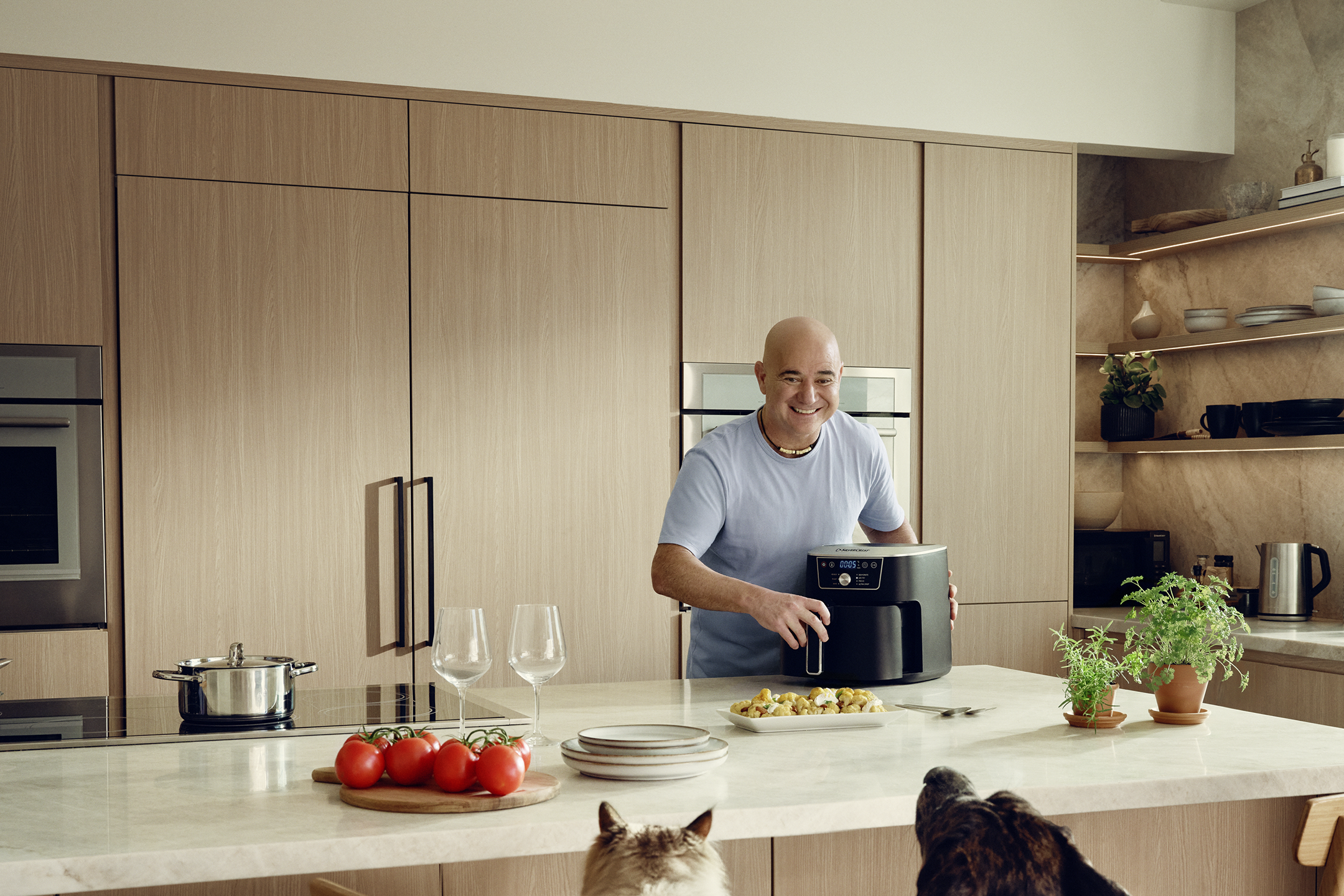 Andre Agassi în bucătărie cu o friteuză cu aer cald, preparând mâncare în timp ce câinii privesc.
