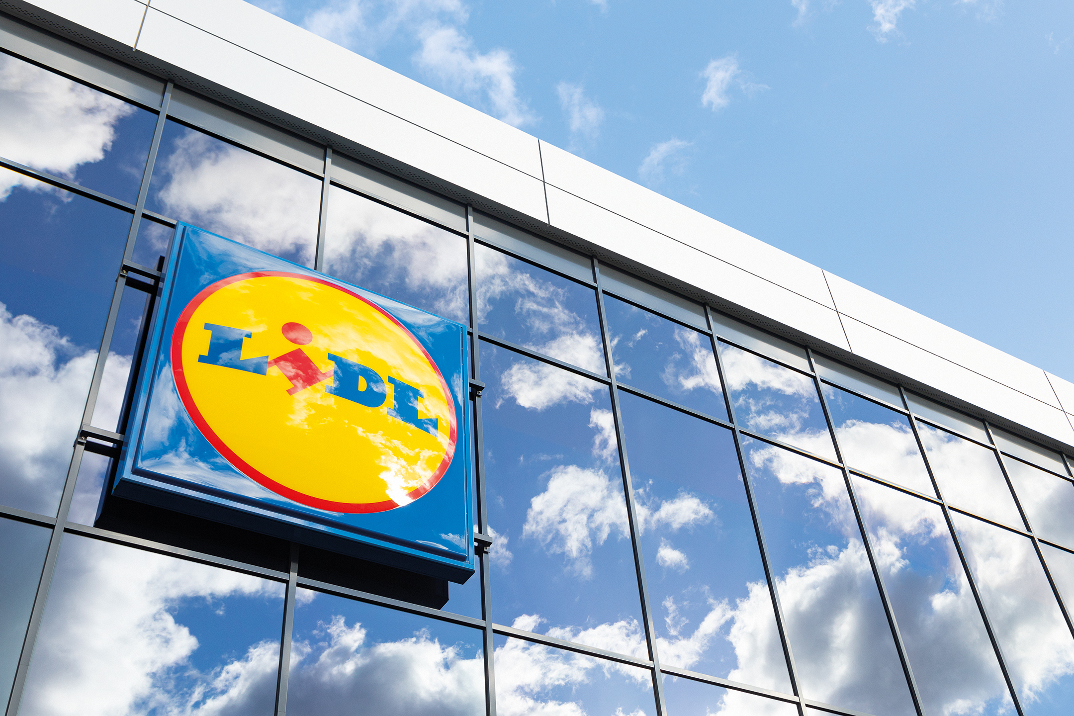 Semnul Lidl pe o fațadă de sticlă a unei clădiri, cu cerul și norii reflectați.