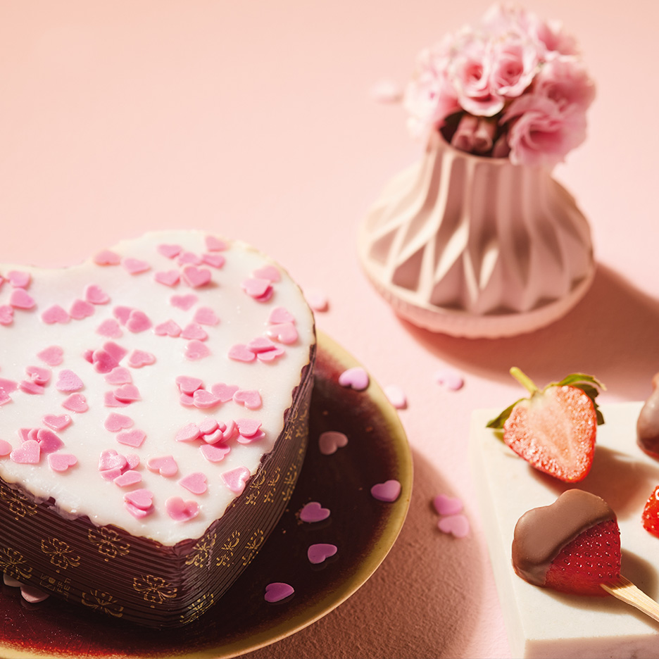 Tort în formă de inimă cu glazură albă și inimioare roz, flori și căpșuni cu ciocolată.