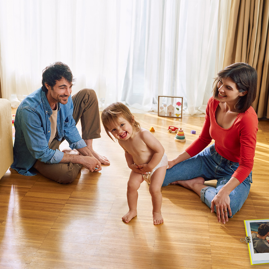 Părinți zâmbitori se joacă cu bebelușul lor fericit, care poartă un scutec, pe podeaua din lemn.