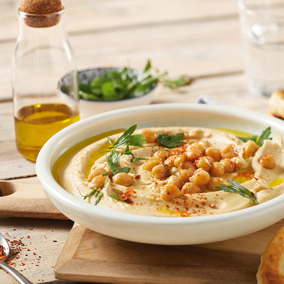 Hummus cu năut, ulei de măsline și pătrunjel, servit într-un bol alb pe o masă de lemn.