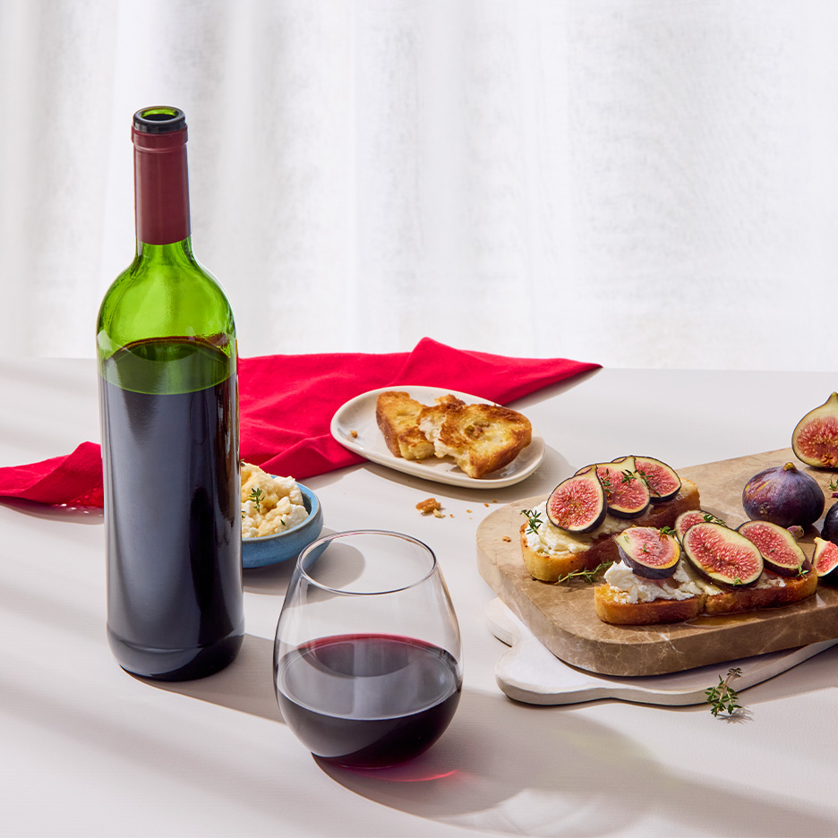 O sticlă de vin roșu, un pahar cu vin și bruschete cu smochine și brânză pe o masă.