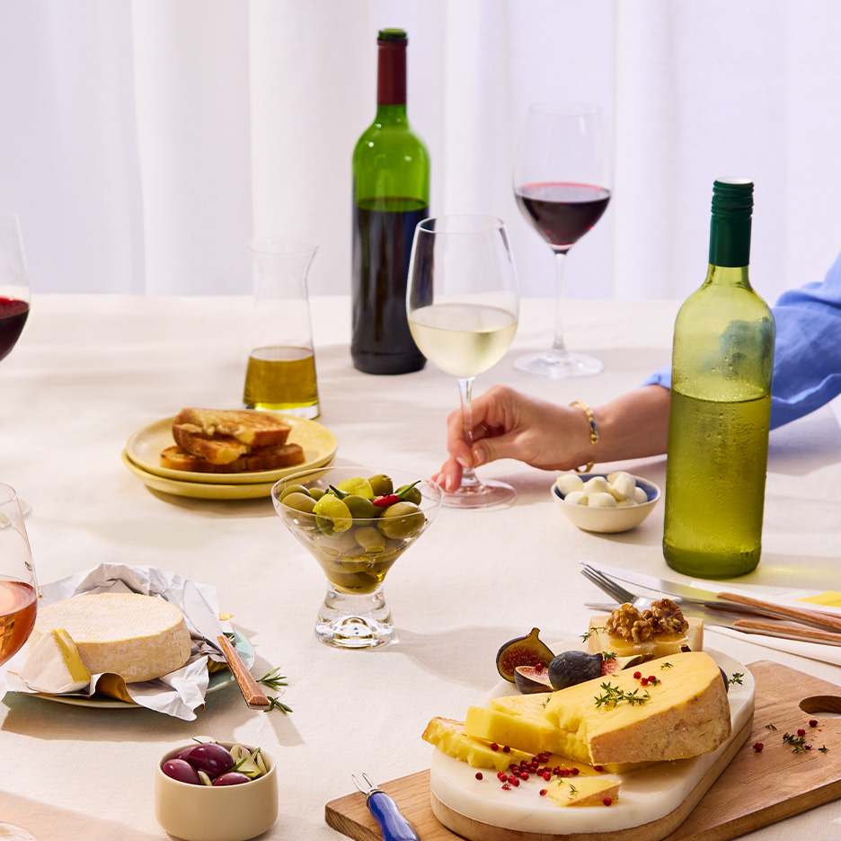 Masă cu vin, brânzeturi, măsline și pâine prăjită, cu o persoană care ține un pahar de vin.