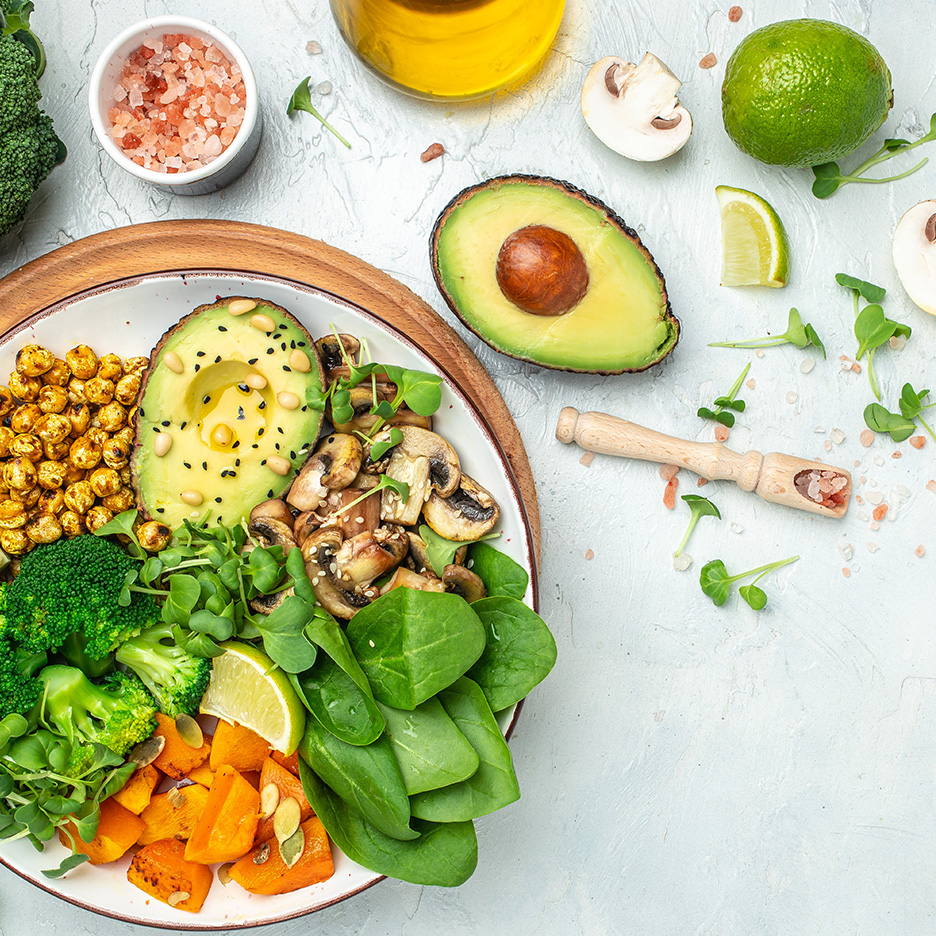 Bol vegan cu avocado, năut, broccoli, ciuperci, spanac și dovleac, pe o masă deschisă.