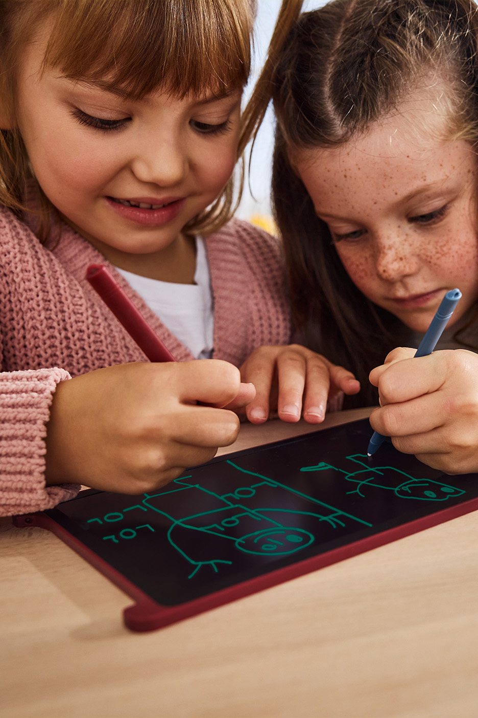 Două fetițe desenează pe o tabletă de scris cu stilouri roșu și albastru.