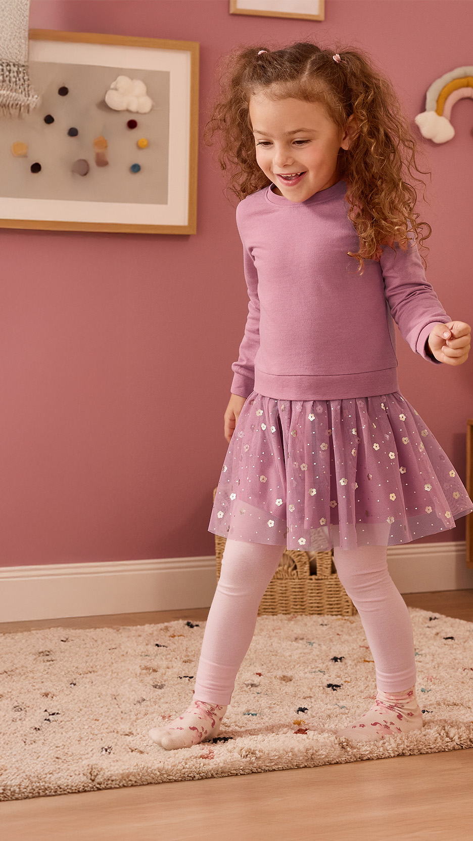 Fetiță zâmbitoare în bluză și fustă mov, colanți roz, pe un covor pufos.