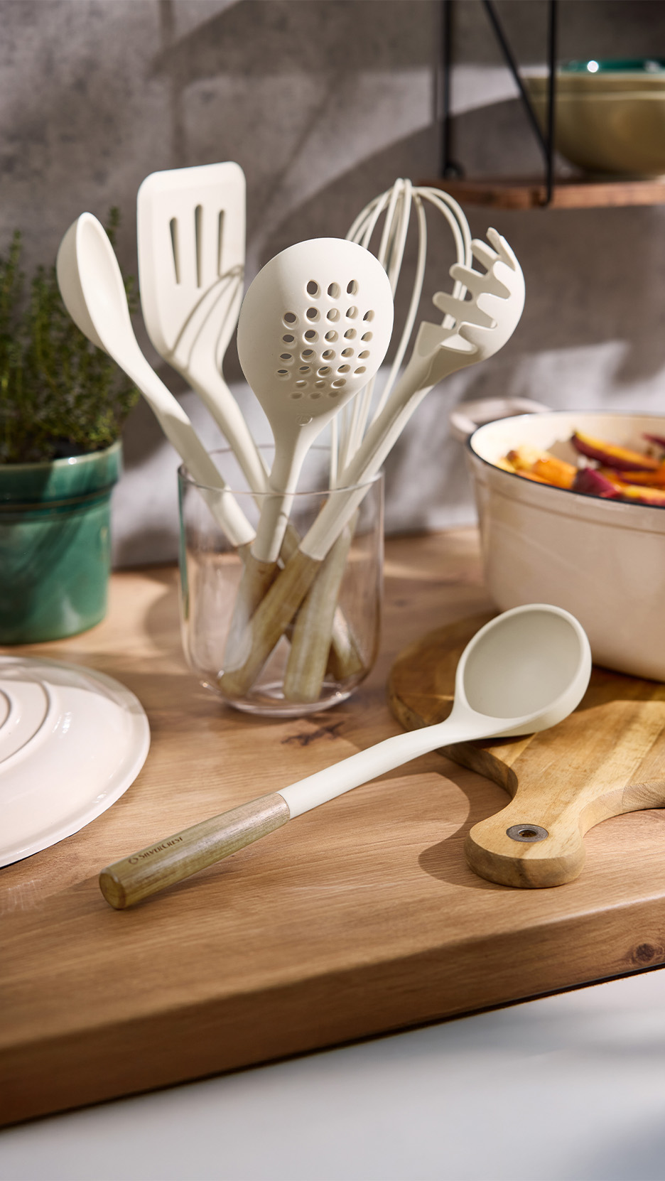 Set de ustensile de bucătărie crem cu mânere din lemn, inclusiv o spatulă, polonic și tel, pe o masă de lemn.