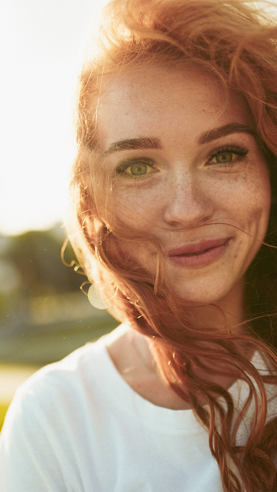 Femeie zâmbitoare cu păr roșcat și pistrui, purtând un tricou alb, în lumină caldă.