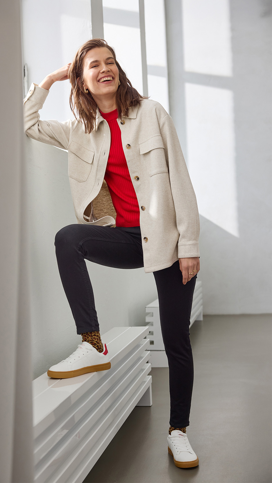 Femeie zâmbitoare în jachetă bej, pulover roșu, pantaloni negri și adidași albi cu șosete leopard.