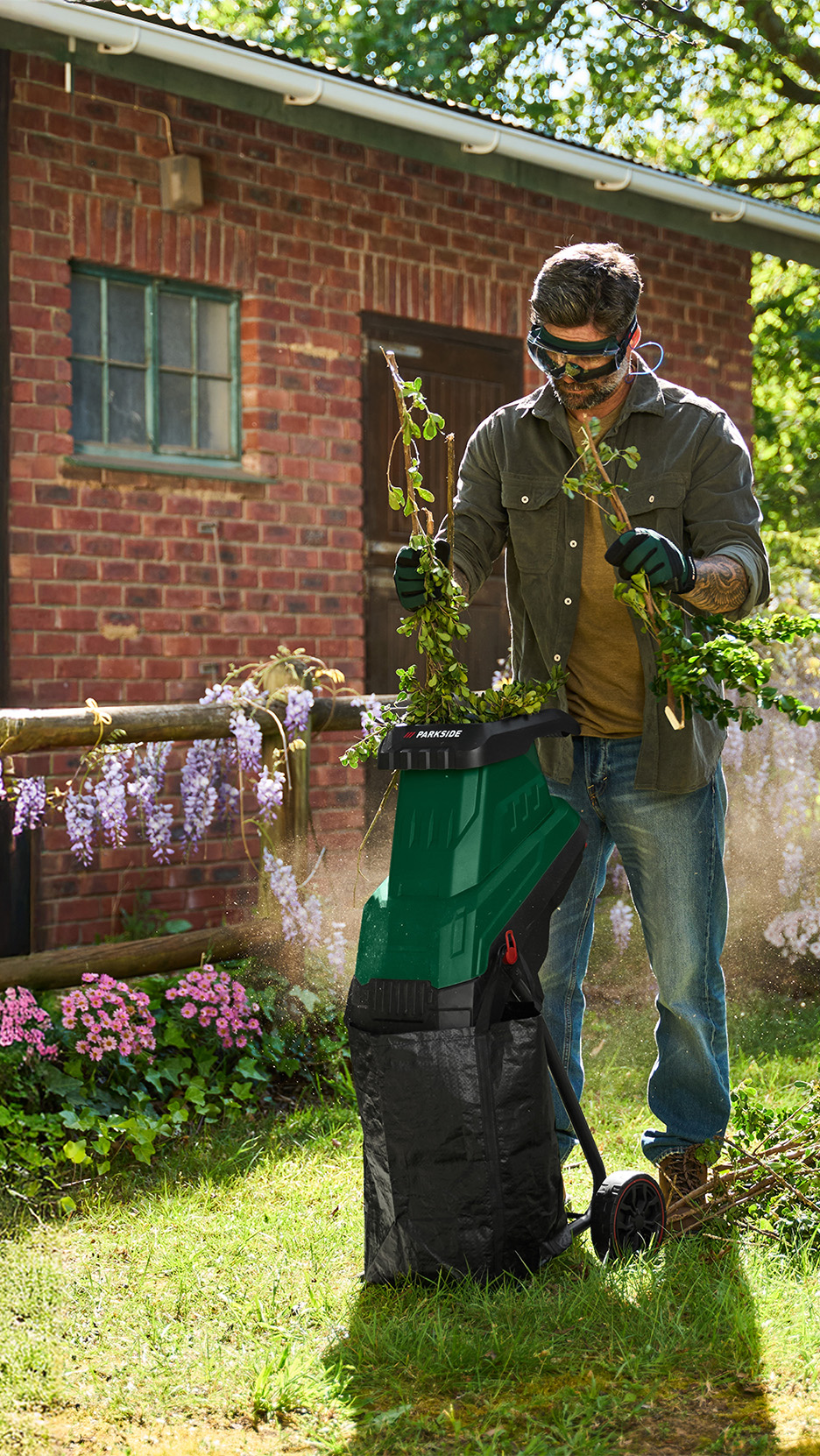 Bărbat cu ochelari de protecție și mănuși, folosind un tocător de grădină verde pentru a mărunți crengi.