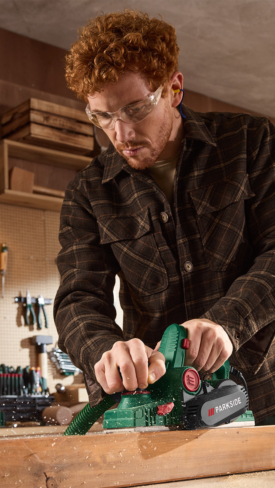 Bărbat cu ochelari de protecție și căști de urechi, folosind o rindea electrică pe o scândură de lemn.