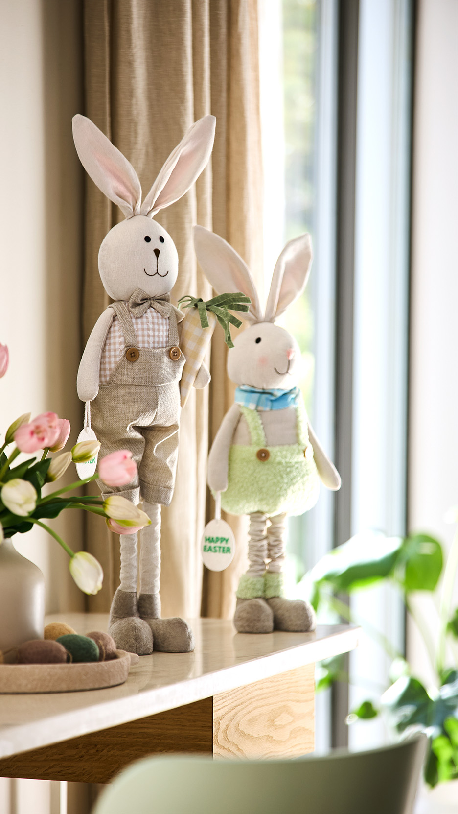 Două figurine iepurași de Paște pe o masă, cu flori și ouă decorative.