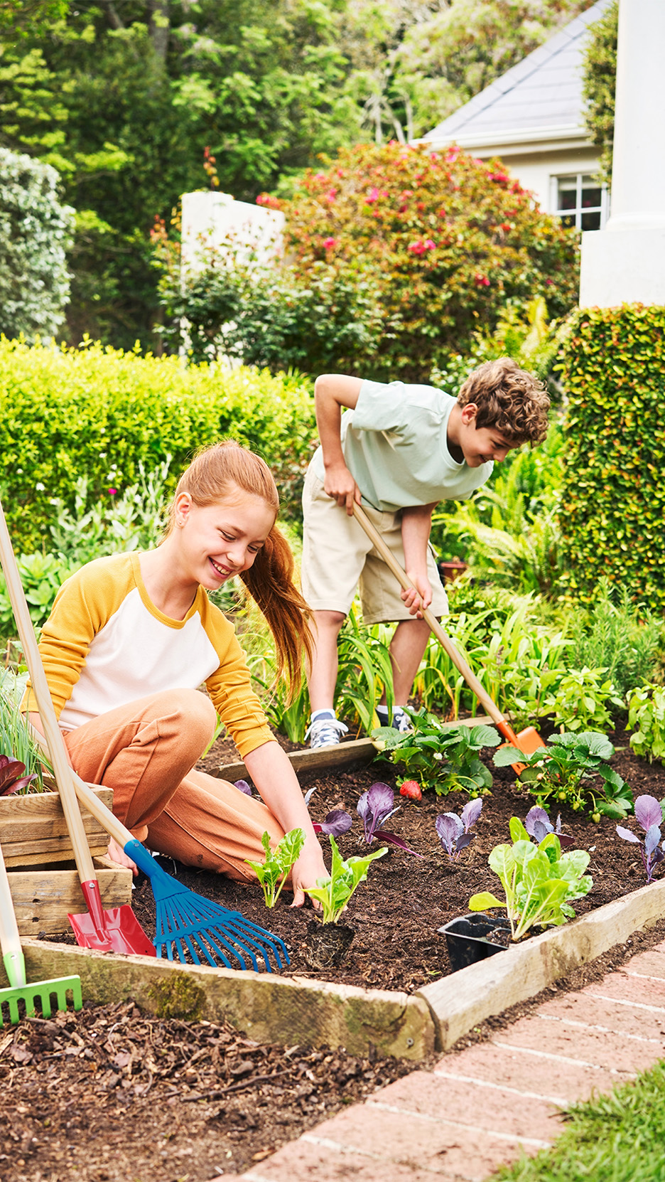 Doi copii zâmbitori plantează răsaduri într-o grădină, folosind unelte de grădinărit.