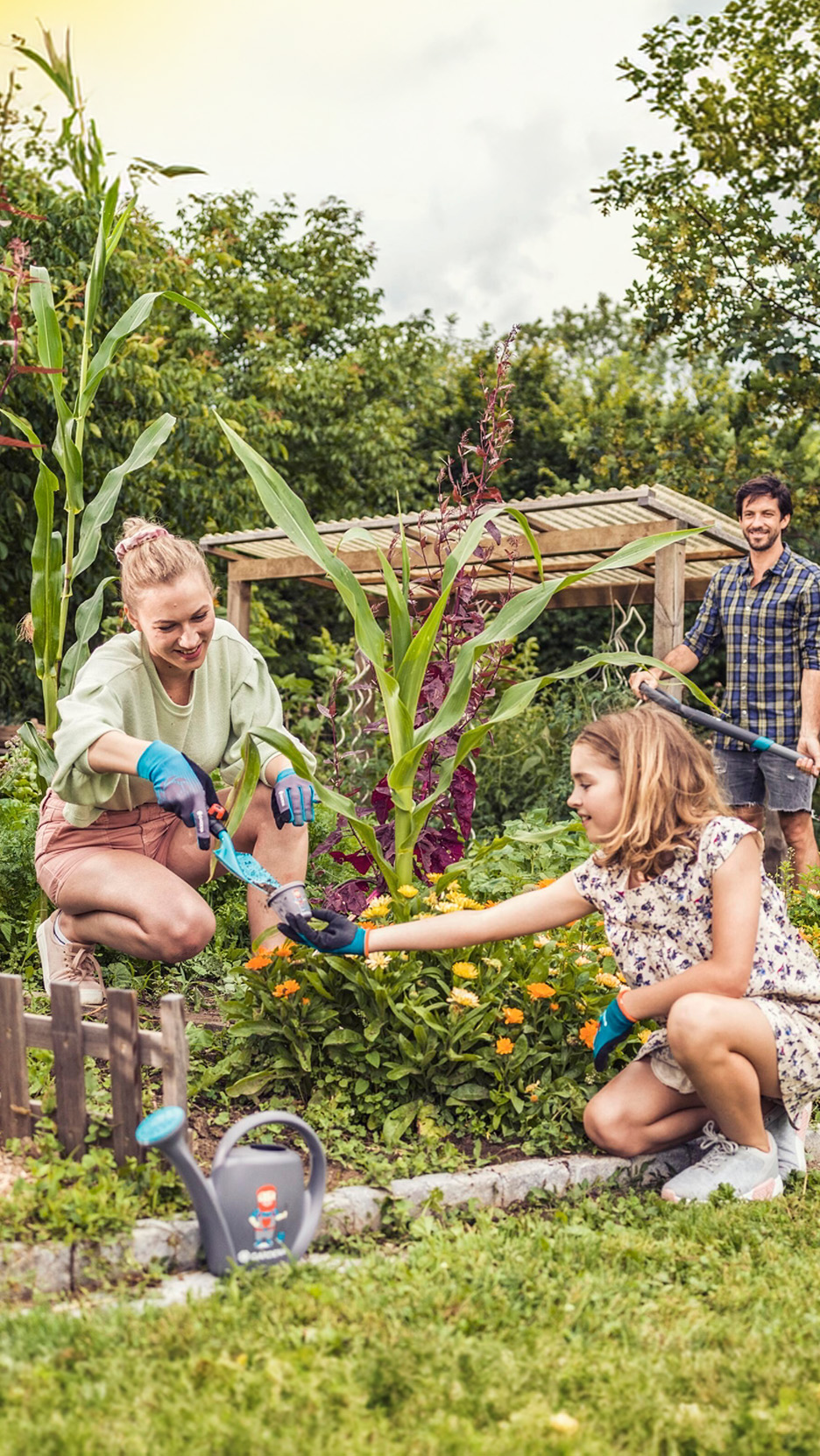 Familie grădinărind, cu o femeie și o fată plantând flori, iar un bărbat udând plantele.
