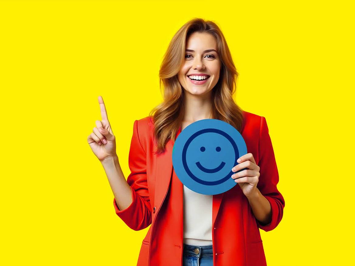 Femeie zâmbitoare în roșu, ținând un emoticon fericit și arătând în sus.