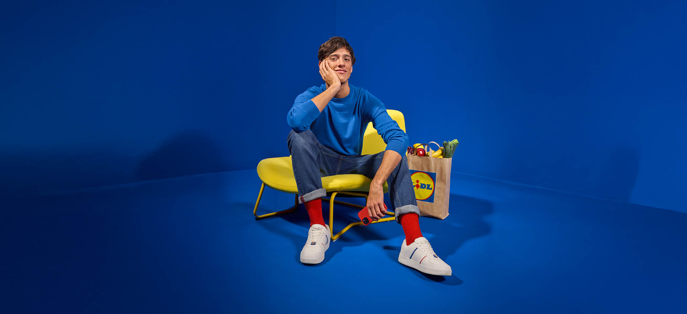 Bărbat așezat pe un scaun galben, cu o pungă de cumpărături Lidl plină cu fructe și legume.