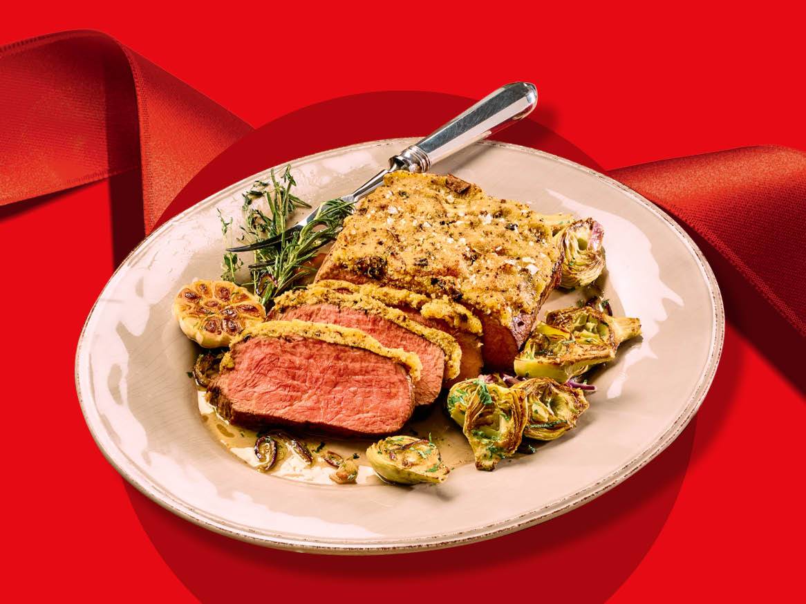 Friptură de vită cu crustă, anghinare și usturoi pe o farfurie, cu o panglică roșie în fundal.