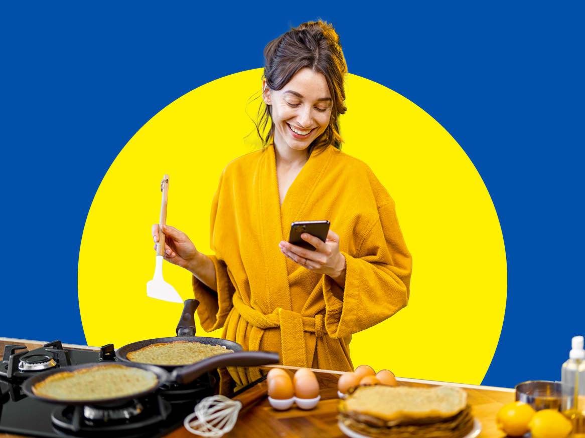 Femeie în halat galben, făcând clătite și uitându-se la telefon, pe fundal albastru și galben.