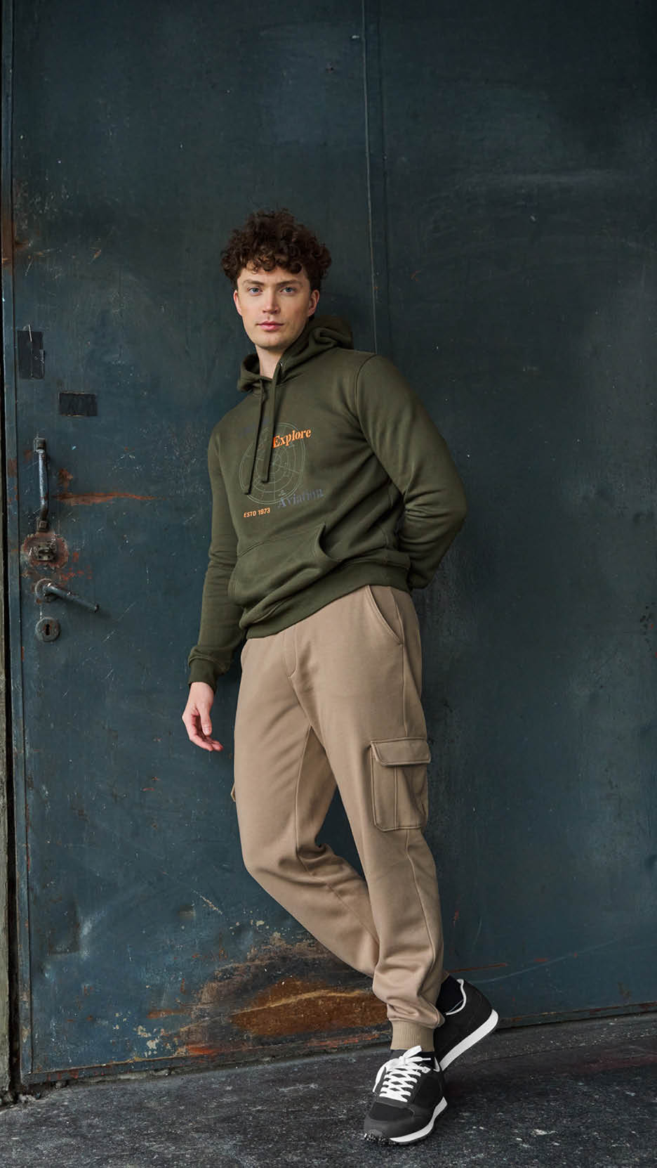 Bărbat într-un hanorac verde kaki cu imprimeu 'Explore' și pantaloni cargo bej.