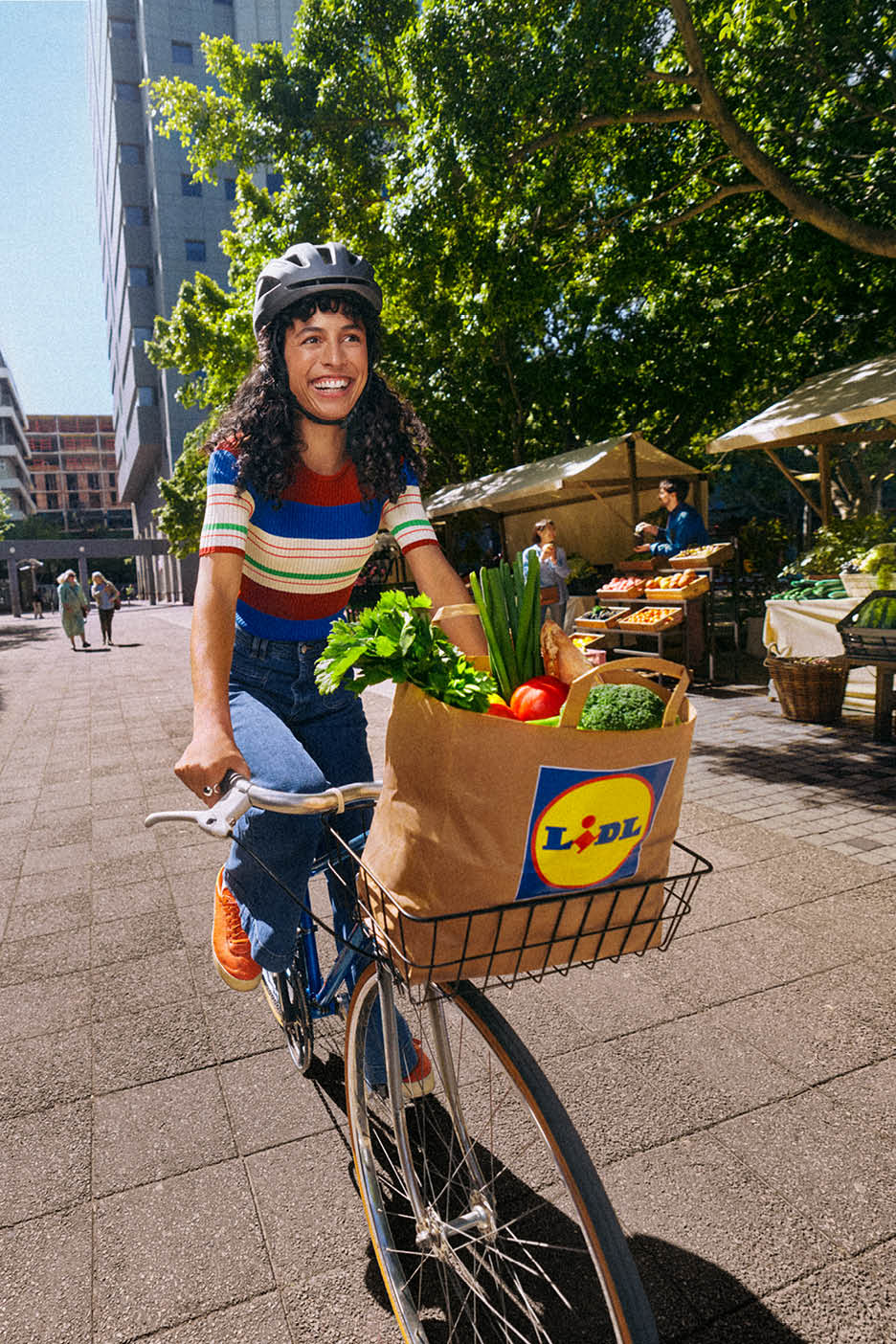 Femeie zâmbitoare pe bicicletă, cu un coș plin de legume proaspete și produse alimentare.