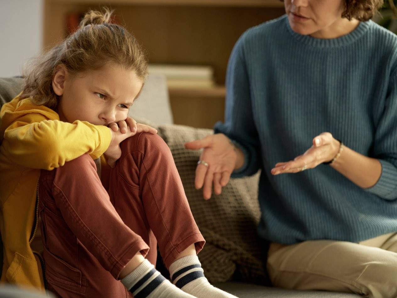 O mamă și fiica ei pe o canapea, fiica fiind supărată și mama gesticulând.